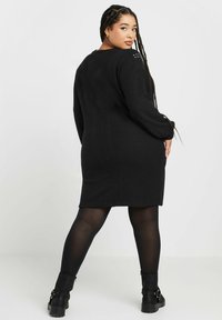 Abito maglione nero con maniche lunghe, vestibilità comoda, tessuto testurizzato e borchie decorative sulle spalle, abbinato a collant neri e stivaletti alla caviglia.