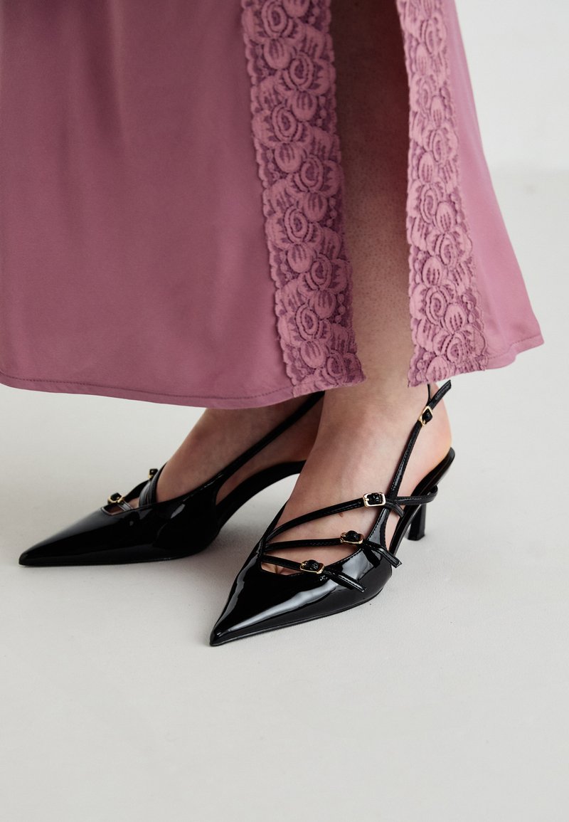 Tacchi slingback in vernice nera con punta affusolata, caratterizzati da numerose cinghie sottili e dettagli in oro.