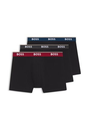 Muskeløs mørkhudet mand står med hænderne bag ryggen iført sorte BOSS-brandede boxertrusser mod en ensfarvet hvid baggrund.