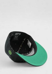 Musta lakki, jossa vihreä lippa, logokoriste ja sisävuoren yksityiskohtia. Materiaali vaikuttaa teksturoidulta, ja lippiosa on kaareva.