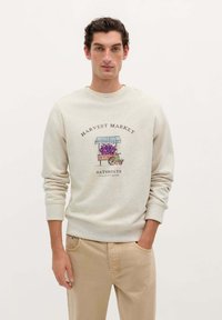 Sudadera gris clara con cuello redondo, que presenta un diseño ilustrado de un mercado de cosecha con uvas y un puesto. Combinada con pantalones beige.
