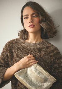 Metallic-goldene Clutch-Tasche mit strukturierter Oberfläche, gehalten von einer Person mit braunem, flauschigem Pullover. Weiches Licht hebt Details hervor.