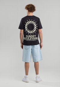 Musta puuvillapaita, jossa on auringon grafiikka ja "SUNSET BOULEVARD" -teksti vaaleassa värissä, yhdistettynä vaaleansinisiin farkkushortseihin ja valkoisiin tennareihin.