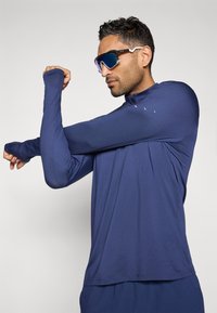 Μπλούζα αθλητική με μακριά μανίκια σε ναυτικό μπλε, κατασκευασμένη από ελαφρύ ύφασμα με γιακά τύπου μισό φερμουάρ. Διαθέτει τρύπες για τον αντίχειρα και εφαρμοστή σχεδίαση για ευκολία στην κίνηση.