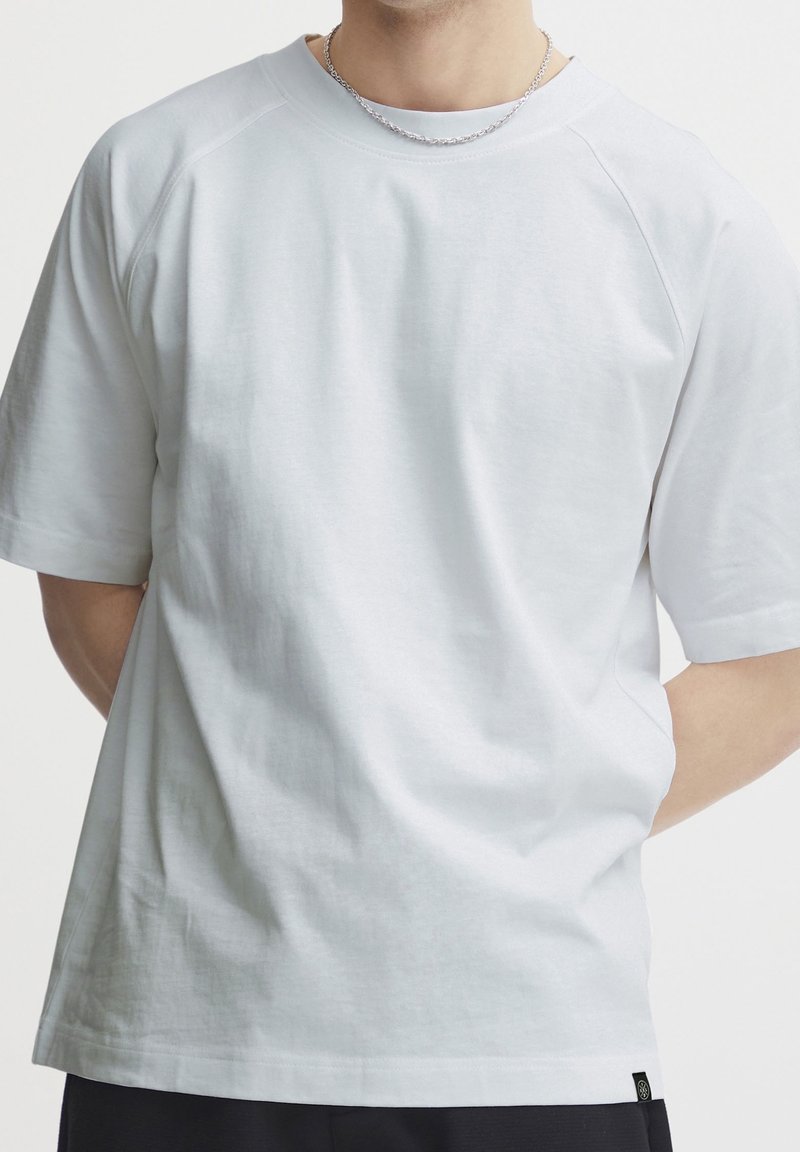 Homme portant un t-shirt blanc simple à manches courtes et un collier en chaîne argentée, les mains derrière le dos sur fond blanc.