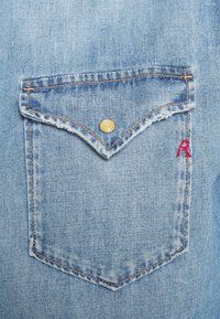 Ljust blå denimtyg har en bröstficka med tvåfärgad söm och en gul tryckknapp, prydd med ett litet rödbroderat "R".