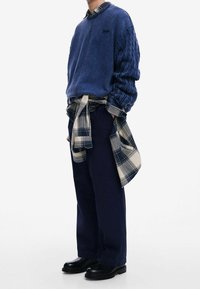 Sweat-shirt bleu avec des manches en maille texturée, pantalon bleu marine, et une chemise à carreaux nouée autour de la taille. Complété par des bottes noires.