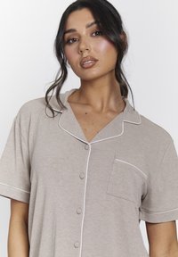 Camisa beige de botones con mangas cortas, hecha de tejido acanalado. Presenta ribetes blancos y un bolsillo en el lado izquierdo.