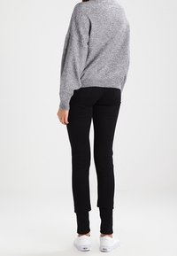 Une femme se tient de dos face à la caméra, portant un pull gris, un pantalon noir et des chaussures blanches. L'arrière-plan est simple et clair.