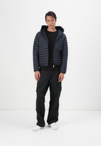 Navy puffer jacket with horizontal quilting, attached hood, front zipper, and orange pull tab. Paired with black cargo pants and white sneakers.