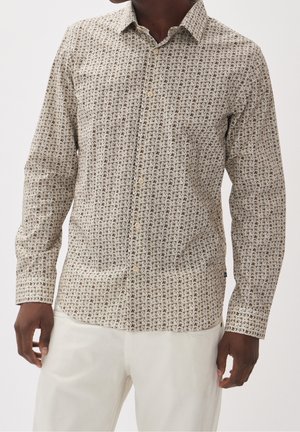 Mann trägt beige langärmliges Hemd mit kleinem braunem und schwarzem Palmenmuster und off-white Hose vor einfarbigem Hintergrund.