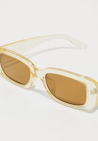 Gafas de sol transparentes con montura amarillo claro y lentes marrones. Forma rectangular con ligera curvatura y patillas transparentes.