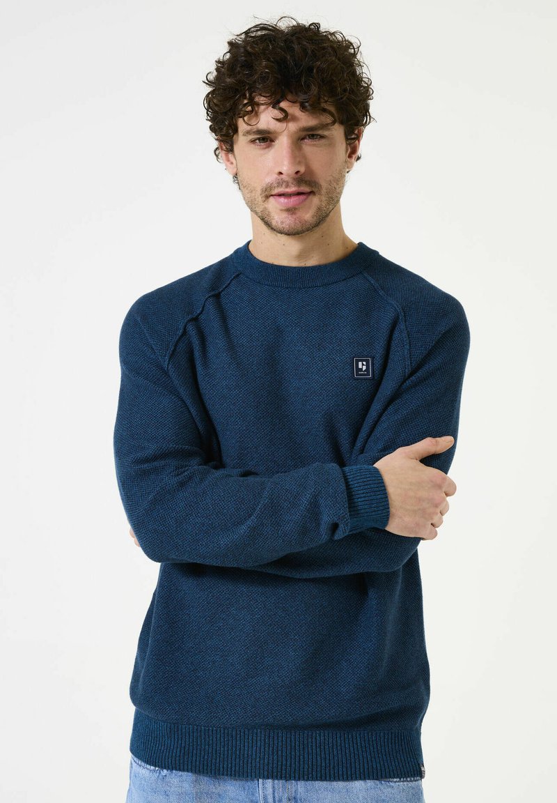 Pull col rond bleu marine avec un motif texturé, poignets et ourlet côtelés. Présente un petit logo en patch sur la poitrine. Tissu tricoté doux.