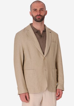 Uomo con la barba che indossa un blazer beige sopra una camicia marrone con collo e pantaloni chiari, in piedi su uno sfondo semplice.