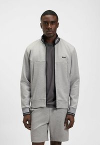 Felpa zip grigia chiara con strisce nere e logo, indossata sopra una maglietta grigia scura e abbinata a pantaloni corti dello stesso colore.