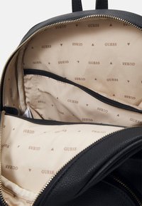Zaino nero con un'esterno testurizzato e zip dorata. L'interno presenta una fodera beige con il logo "GUESS" ripetuto e motivi triangolari.