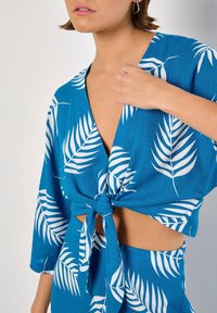 Top corto tropical blu con motivi di foglie bianche, maniche corte e dettaglio con nodo frontale, realizzato in un tessuto leggero e testurizzato.