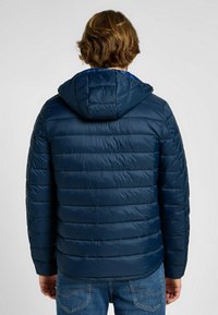 Tamno plavi puffer jakna s horizontalnim kvadratima, kapuljačom i laganom, sjajnom tkaninom; u kombinaciji s plavim trapericama.