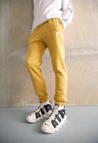 Gula joggers i bomull med elastiska muddar och en vit Nike-logotyp, kombinerade med vita och svarta sneakers med tydliga metallaccenter.