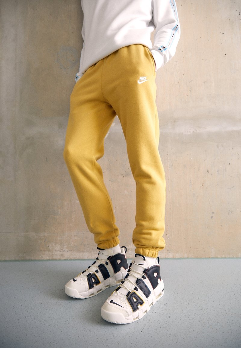 Gula joggers i bomull med elastiska muddar och en vit Nike-logotyp, kombinerade med vita och svarta sneakers med tydliga metallaccenter.