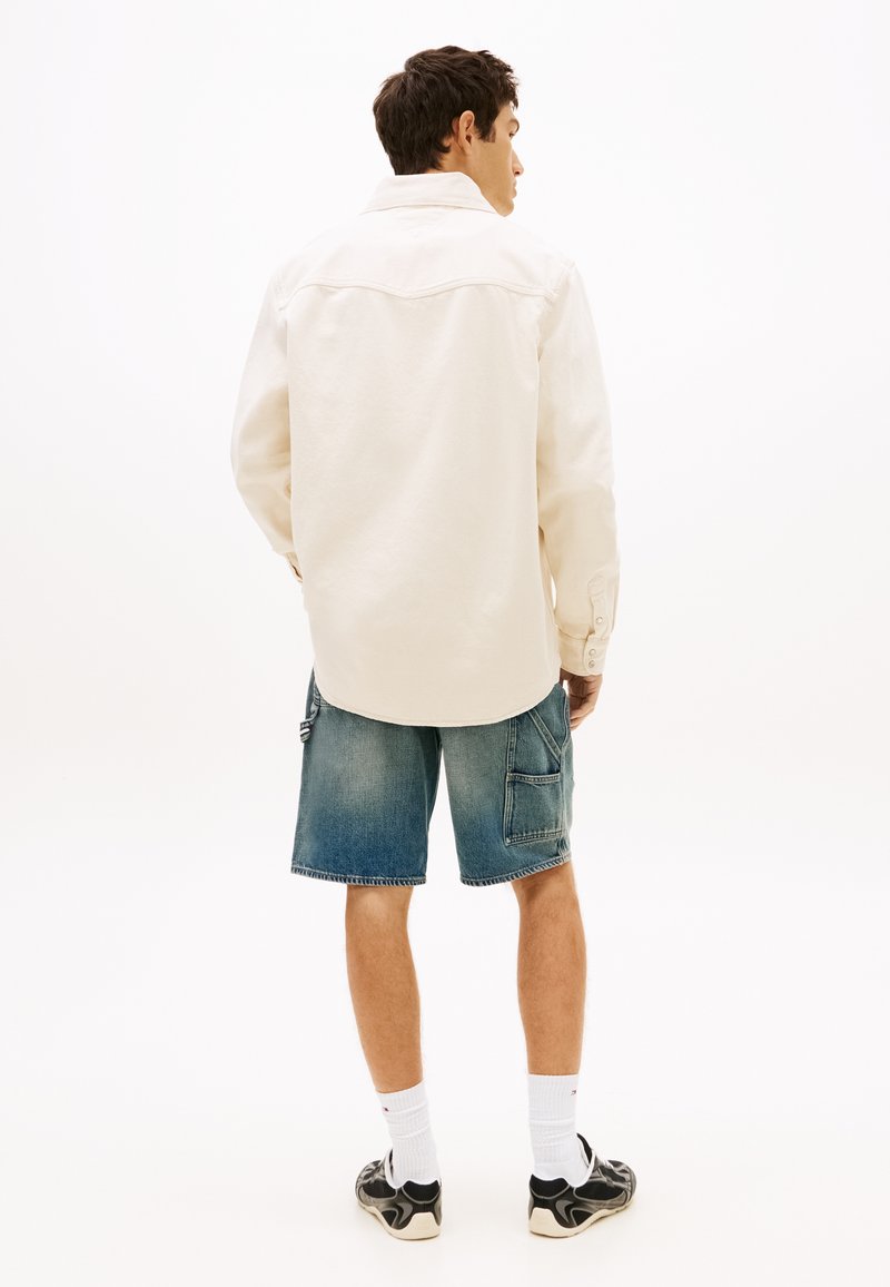 Man die wegkijkt, draagt een crème-kleurig overhemd met lange mouwen, denim cargoshorts, witte sokken en zwarte sneakers tegen een witte achtergrond.