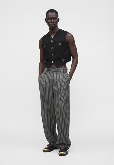 Homme debout portant un gilet noir sans manches avec des boutons dorés et un pantalon large rayé avec des chaussures noires à bouts dorés.