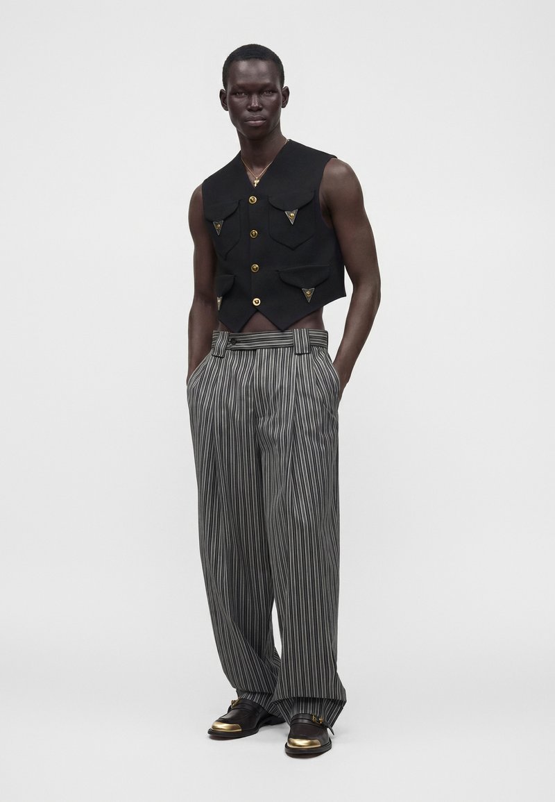Homme debout portant un gilet noir sans manches avec des boutons dorés et un pantalon large rayé avec des chaussures noires à bouts dorés.