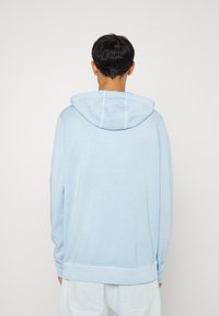 Sudadera con capucha de color azul claro, de corte relajado, textura suave, con puños y dobladillo acanalados. Cuenta con un bolsillo tipo canguro y capucha ajustable con cordón.