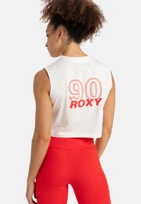White cropped tank top with orange "90 Roxy" graphic on the back, paired with red high-waisted leggings. Soft cotton material, sleeveless design.