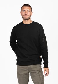 Sudadera negra con textura acanalada y escote redondo. Presenta un bolsillo con cremallera en la manga izquierda. Se combina con pantalones cargo color caqui.