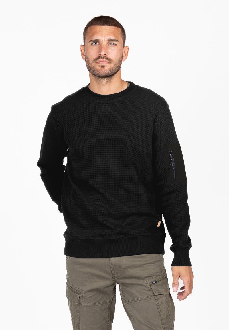 Sudadera negra con textura acanalada y escote redondo. Presenta un bolsillo con cremallera en la manga izquierda. Se combina con pantalones cargo color caqui.