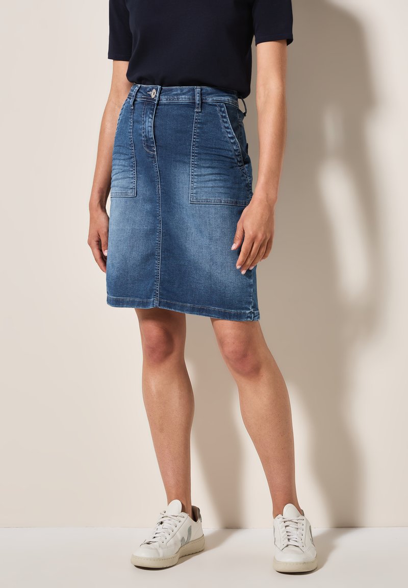 Jupe en denim bleu clair, longueur genoux avec poches avant, passants de ceinture et détails de texture subtils. Assortie avec des baskets blanches.
