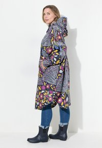 Manteau imperméable coloré avec des motifs floraux et géométriques, capuche et poches latérales, porté sur un jean bleu et des bottines noires.