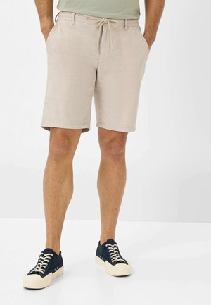 Pantaloni corti di lino beige con cintura elastica e cordoncino, dotati di tasche laterali. Abbinati a sneaker in tela blu navy con suole bianche.