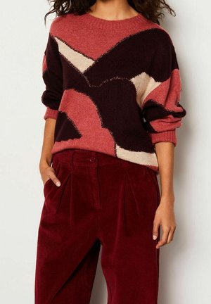 Donna che indossa un maglione con motivo a forme bordeaux, rosa e crema e pantaloni rossi in velluto a coste a vita alta con le mani nelle tasche.
