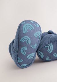 Chaussons bleus à texture douce, présentant un motif de petits arcs-en-ciel bleu sarcelle. La semelle est ajustée avec des formes arrondies.