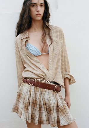 Kobieta o długich ciemnych włosach nosi jasnoniebieski top bikini pod beżową koszulą w kratę, zestawiony z dopasowaną plisowaną spódnicą i szerokim brązowym paskiem.