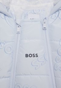 Světle modrá péřová bunda s měkkým bílým vnitřkem, s černým logem „BOSS“ a jemnými reliéfními vzory, dvojitými zipy a žebrovanými manžetami.