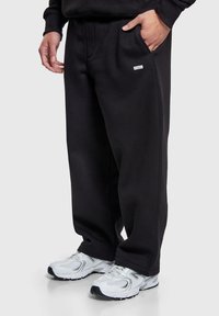 Pantalon de survêtement en coton noir avec poches latérales et petit détail logo ; associé à des chaussures de sport blanches avec des accents gris et des semelles texturées.
