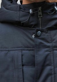 Schwarze isolierte Jacke mit hohem Kragen, durchgehendem Reißverschluss, verstellbarer Kordelzugkapuze und Knopfdetails auf der Frontpartie.