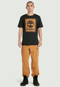 Czarna koszulka z krótkim rękawem z pomarańczową grafiką Timberland, zestawiona z beżowymi spodniami cargo i czarnymi butami. Prosty, codzienny strój.