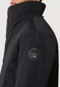 Personne portant une veste noire à col montant avec un écusson circulaire sur le haut du bras représentant un globe et du texte.