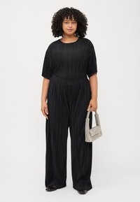A woman with curly hair stands wearing a black pleated jumpsuit, holding a small, textured handbag against a plain white background.