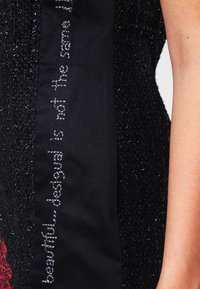 Svart texturerad tyg med en slät svart remsa som har silverbroderad text som lyder: "vacker...desigual är inte detsamma."