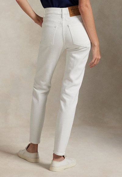 Persoon in witte high-waisted slim jeans met achterzakken en witte sneakers, staande tegen een neutrale achtergrond.