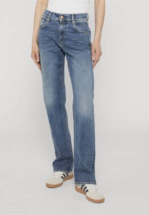 Personne portant un jean bleu clair droit et des baskets blanches avec des rayures noires, debout devant un fond clair uni.