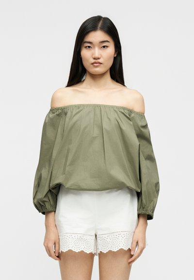 Γυναίκα που φοράει μπλούζα λαδί με ανοιχτούς ώμους και φουσκωτά μανίκια, καθώς και λευκά σορτς με δαντέλα eyelet, στέκεται μπροστά σε απλό φόντο.