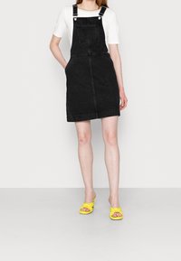Black denim overall dress with adjustable straps, front pocket, and a straight-cut shape. Worn over a white shirt with yellow heeled sandals.