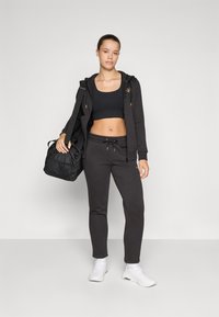 Zwart sportoutfit bestaande uit een aansluitende crop top en sweatpants, gecombineerd met een vest met rits en witte sneakers. Een zwarte tas hangt van de schouder.