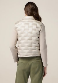 Vesta pufo crem cu model matlasat, guler înalt și finisaj neted. Asortată cu un top bej deschis cu mânecă lungă și pantaloni verzi măslinii.